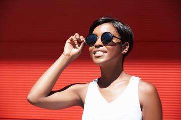 Young stylish charismatic African woman trying her new contemporary fashionable round-shaped sunglasses, looking at the sun, having positive face expression, isolated against red wall background