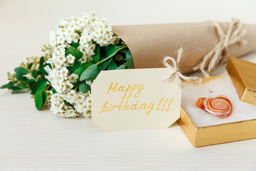 Fototapeta na wymiar Birthday Card with Golden Present Box with Glass Heart. Bouquet White Small Flowers in Brown Craft Paper with String.White Wooden Texture Background.Selective Focus