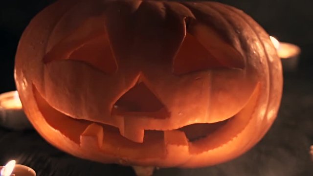 Carved Halloween pumpkin lit a flame inside, around the smoke travel along on a black background with lighted candles close up. Slowmotion
