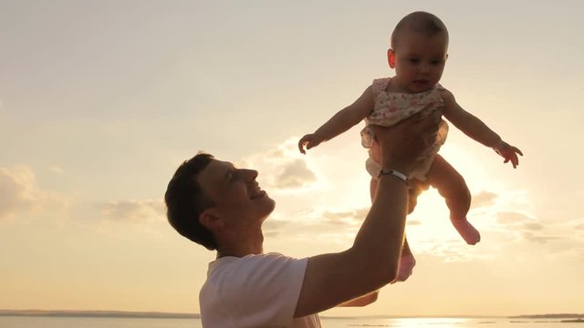 Happy Man Playing Lifting His Daughter Outdoors. Silhouette Of Young Father Holding Up His Cute Baby Daughter In Air Against Sunset Sky Playfully