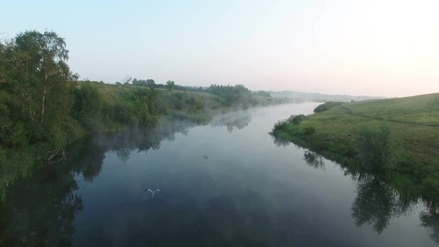 Aerial video of a big lake at sunrise while fog creeping over the water. Cranes fly over the lake. Drone moving forward. 4K Aerial stock footage shot at summer season time.
