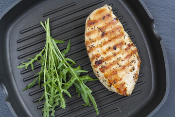 Grilled chicken breast steak with arugula rucola on teflon pan grill, black slate stone background