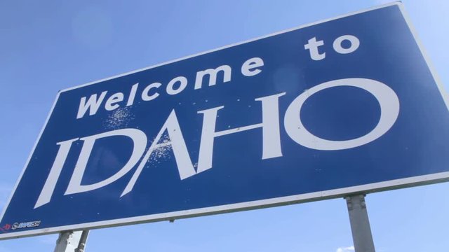 Welcome To Idaho Sign, Pan. Low angle pan to and from a blue "Welcome To Idaho" sign placed along the highway.