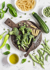 Raw ingredients for cooking vegetarian lunch - dry pasta, fresh broccoli, zucchini, green peas and beans,  olive oil. On a light background, top view
