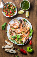 Plate of roasted tiger prawns and pieces of octopus with fresh leek, vegetable salad, peppers, lemon, bread and pesto sauce over wooden background, top view, vertical composition