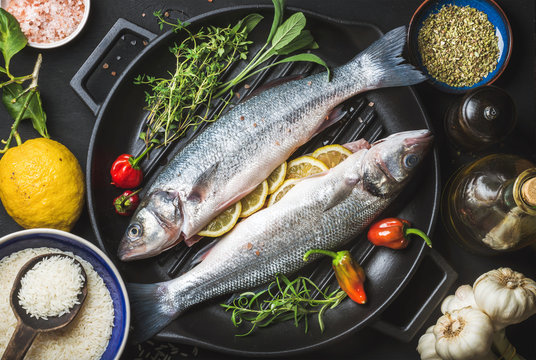 Ingredients for cookig healthy fish dinner. Raw uncooked seabass fish with rice, lemon, herbs and spices on black grilling iron pan over dark background, top view, horizontal composition