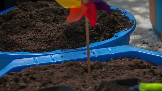 Small garden tools are placed on a soil in a big container. A colourful paper windmill is inserted in there, too. Close-up shot.
