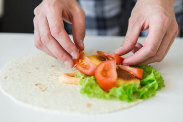 man prepares to roll fast food.
chicken nuggets, lettuce, tomato, cheese, Mexican tortillas