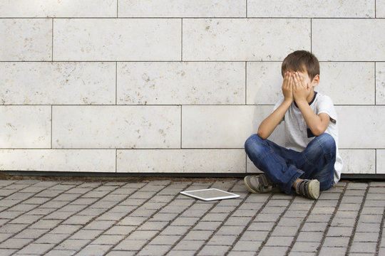 Sad, lonely, unhappy, disappointed child sitting alone on the ground. Boy holding his head, look down. The tablet pc computer near him. Casual clothes. Outdoor