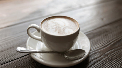 Cup of tasty latte is standing on the wooden textured table. There is nice picture is painted on the top of the latte. Latte is in the white big cup.