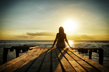 Fototapeta premium Girl in denim dress sitting on a pier on the beach and looking at the water and sun. Meditation and relaxation.