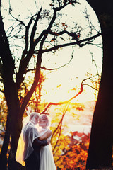 Newlyweds hidden under a veil stand in the gold autumn forest