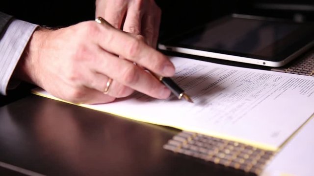 Businessman sitting office desk signs a bill comparing them, with the pc tablet. close-up hands of a man and a fountain pen