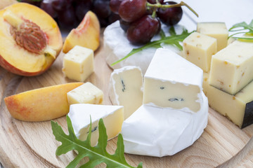 Different delicious cheeses camembert, blue cheese, cheese with nut, fruits red grapes and peach and herbs mint and arugula on wooden round board