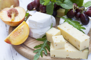 Different delicious cheeses camembert, blue cheese, cheese with nut, fruits red grapes and peach and herbs mint and arugula on wooden round board