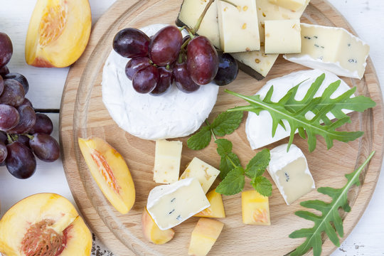 Different delicious cheeses camembert, blue cheese, cheese with nut, fruits red grapes and peach and herbs mint and arugula on wooden round board