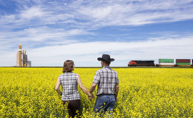 horizontal image of a husband and wife checking canola field while a train rumbles by in the background next to a grain elevator.