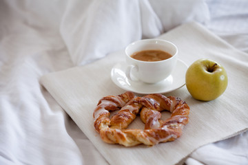 Tray with breakfast on a bed. Sweet pretzel, Cup of coffee and Apple