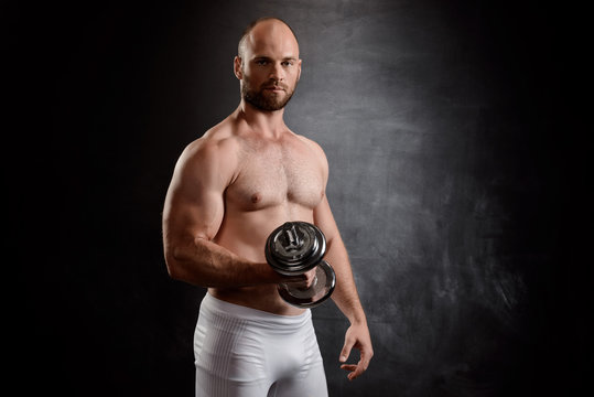 Young powerful sportsman training with dumbbells over black background.