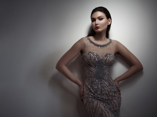 Fatale brunette with expressive dark make up and sleeked hair is posing in the studio on the gray background in the smart beige evening dress embroidered by sequins and beads