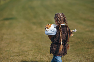 Little girl with pigtails holds a big red cat on a background of an autumn lawn. Back view.