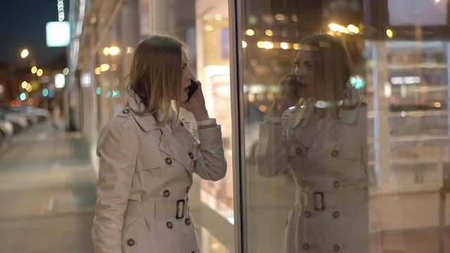 Beautiful woman walking in the street, talking on the cell phone and looking at her reflection in glass store window