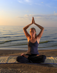 Fototapeta na wymiar Beautiful young woman practising yoga on mat outdoors at river bank on sand at sunset