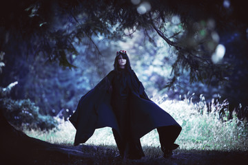 Young black-haired girl in long cloak with a hood in dark forest. Effect of toning - obrazy, fototapety, plakaty