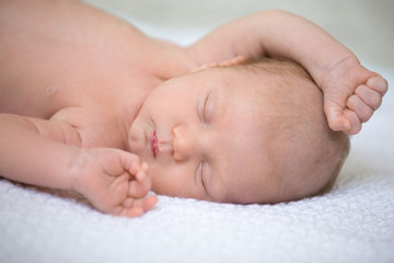 Portrait of young funny newborn babe napping on soft white knitted blanket with his arms up. Cute caucasian new born child sleeping. Healthy one month old kid lying on bed with closed eyes. Close-up