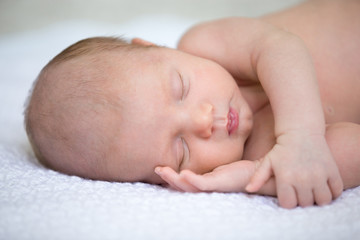 Portrait of young funny newborn babe napping on white knitted blanket with arm under cheek. Cute caucasian new born child sleeping. Healthy one month old kid lying on bed with closed eyes. Close-up