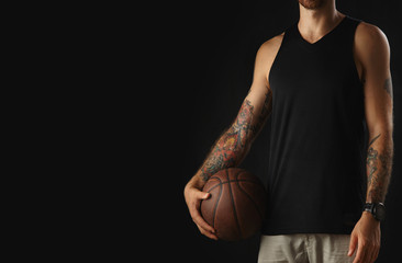 Brutal tattooed athlete in blank sleeveless t-shirt holds leather basketball and ready for game, isolated on black