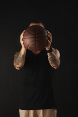 Brutal athlete with tattooed hands in blank sport tank holds focused leather ball for basketball in front on his head, isolated on black