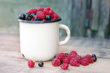 Fresh juicy berries, raspberries, currants, blackberries, a gooseberry in an old white iron mug on a wooden surface