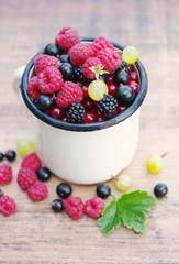 Fresh juicy berries, raspberries, currants, blackberries, a gooseberry in an old white iron mug on a wooden surface