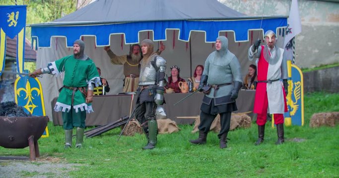 Four knights were kneeling before their king. Now, they stand up and turn around. Two of the men lift their swords up in the air. Wide-angle shot.
