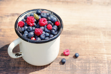 Juicy mature berries of raspberry and bilberry  in an iron mug on a wooden surface.