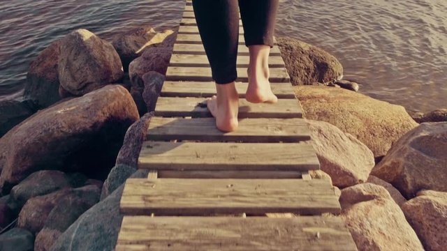 A young barefoot woman is walking out on a small jetty over the ocean in northern Sweden. Camera follows behind legs and feet. Vacation, holiday, recreation concept. Slow motion. Graded look.