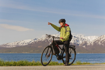 Frau auf E-Bike/ Fahrrad zeigt den Weg. Sie steht mit ihrem Pedelec vor einem Bergpanorama und Wasser. 
Woman on electric bike / bicycle showing the way