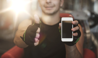 young man with smartphone showing thumbs up in gym