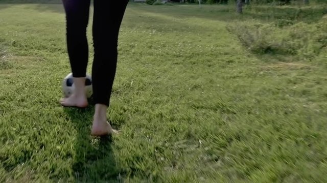Steadicam shot of barefoot girl running with football practicing playing soccer on grass in the summer. Slow motion, camera behind following. Healthy exercise good life concept. Natural look.