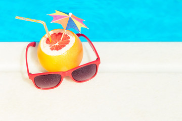 Top down view of grapefruit with paper umbrella and drinking straw and red sunglasses at the edge of swimming pool. Summer vacation symbols. Space for copy. 