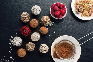 Homemade chocolate and nuts candy balls with cocoa powder, coconut, berries and chopped hazelnuts on black stone background, top view