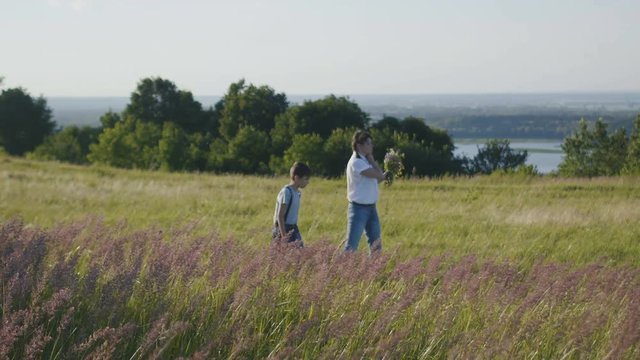 Mother and son in meadow pick flowers for bouquet, summer landscape on high hill