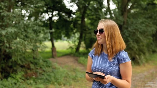 Attractive girl with a tablet in hands walking on the park.
