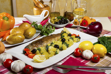 Whole fried trout served on a plate with potato salad and chard with olives, lemon and cherry tomatoes next to fresh vegetables, basil, olive oil and a glass of white wine.