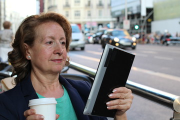 Mature woman with glasses drinking coffee and looking a eletronic tablet using the Internet sitting outdoors in urban cafe. Cafe city lifestyle with traffic lights in the background