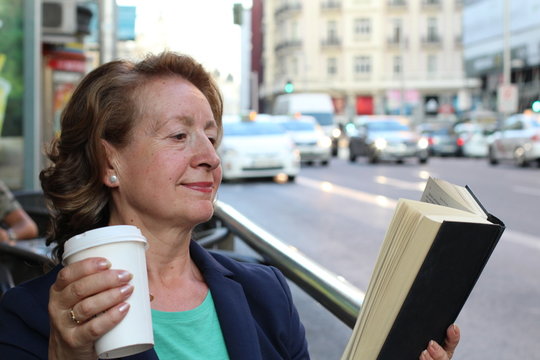 Mature woman drinking coffee and reading book sitting indoor in urban cafe. Cafe city lifestyle with traffic lights in the background