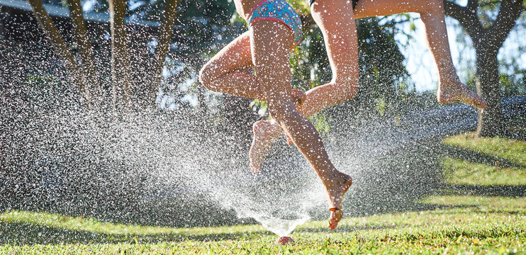 Young girls playing jumping in a garden water lawn sprinkler
