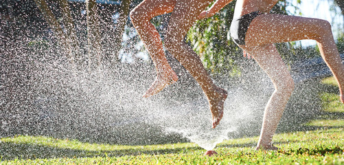 Young girls playing jumping in a garden water lawn sprinkler - obrazy, fototapety, plakaty