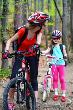 Bikes cycling family. Happy mother and daughter wearing helmet are cycling on bicycle into forest. Mother on bicycle looks back at her daughter.